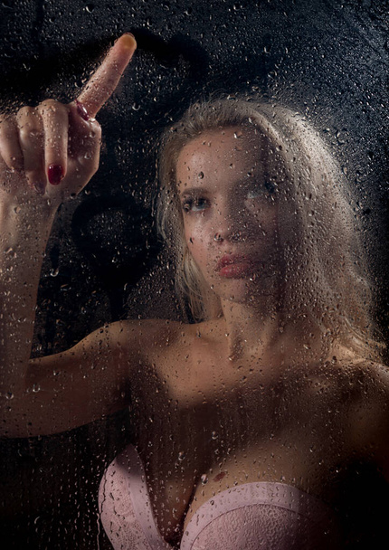 beautyful blonde woman behind wet glass on a dark background, woman takes a shower - Foto, afbeelding