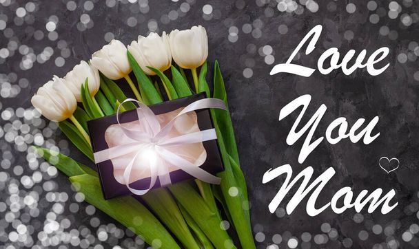 I love You Mom card message sign. Цветы белого тюльпана и черная подарочная коробка на темном фоне с плоскими огнями. Цветочный букет с серебряным бантом, поздравительные открытки вид сверху
. - Фото, изображение