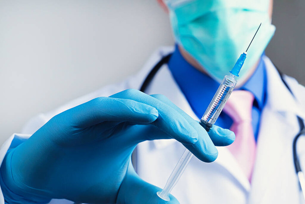 Widok z bliska strzykawki medycznej w gumowej niebieskiej rękawiczce ubranej w dłoń, klatka piersiowa mężczyzny w białej szacie medycznej ze stetoskopem na rozmytym tle - Zdjęcie, obraz
