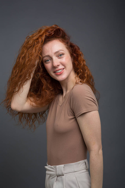 Happy curly red-haired woman with beautiful smile, holding her hair with her hands. On an isolated gray background. Concept: happiness and emotions - Foto, imagen