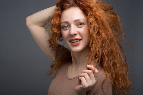 Boldog, göndör, vörös hajú nő, gyönyörű mosollyal, kezében a hajával. Egy elszigetelt szürke háttérrel. Koncepció: boldogság és érzelmek - Fotó, kép
