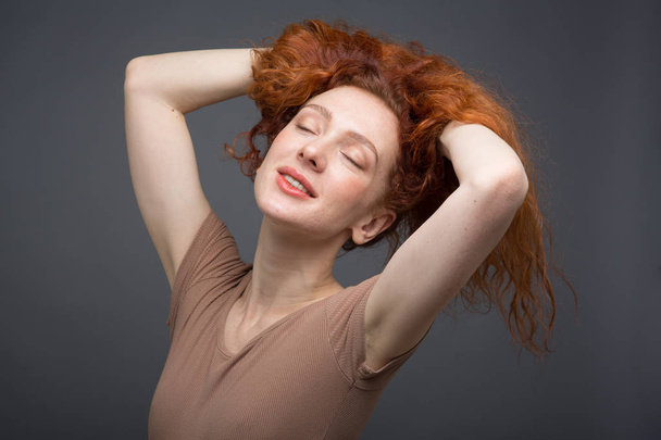 Happy curly red-haired woman with beautiful smile, holding her hair with her hands. On an isolated gray background. Concept: happiness and emotions - Foto, Imagem