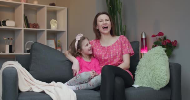 Woman and her little daughter sitting on the sofa and watching comedy film on TV laughing. - Imágenes, Vídeo