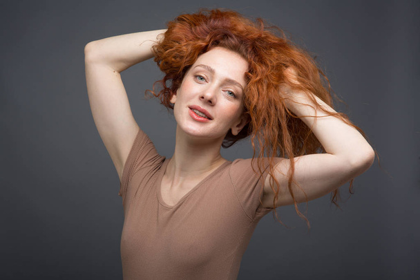 Happy curly red-haired woman with beautiful smile, holding her hair with her hands. On an isolated gray background. Concept: happiness and emotions - Foto, immagini