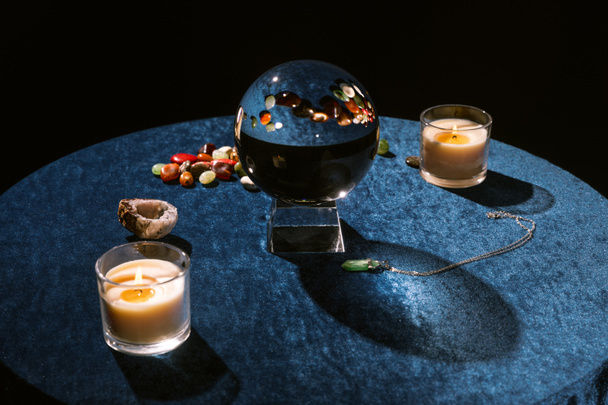 Kristallkugel in der Nähe von Kerzen und Wahrsagersteinen auf dunkelblauem Velourtuch isoliert auf schwarz - Foto, Bild