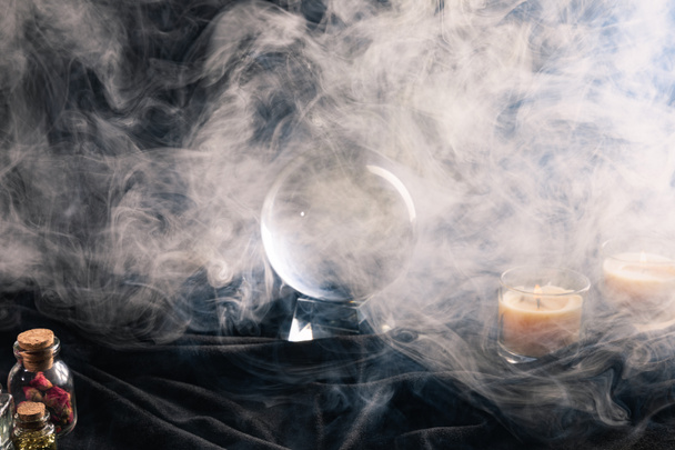 Crystal ball, candles, jars of herbs and buds with smoke around on dark background - Photo, Image