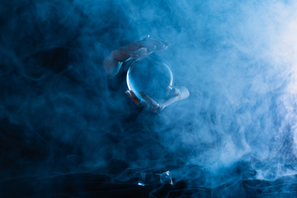 Καλλιεργημένη άποψη της μάγισσας κρατώντας κρυστάλλινη σφαίρα σε σκούρο μπλε φόντο - Φωτογραφία, εικόνα