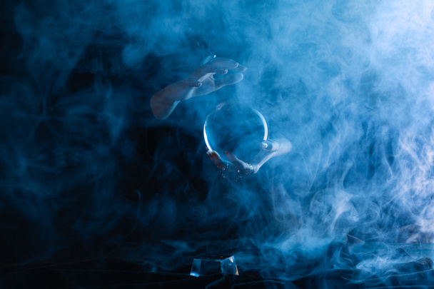 Vista recortada de bruja sosteniendo bola de cristal con humo alrededor en azul oscuro
  - Foto, Imagen