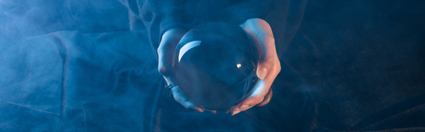 Vista recortada de la bruja sosteniendo la bola de cristal sobre la mesa sobre fondo azul oscuro, tiro panorámico
 - Foto, imagen