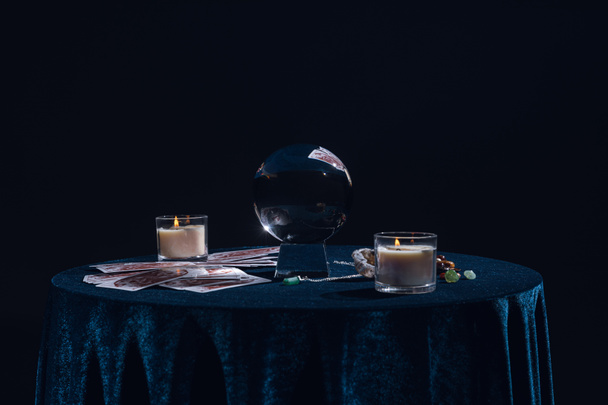 KYIV, UKRAINE - JANUARY 9, 2020: crystal ball with candles, tarot cards, stones and chain on round table isolated on black - 写真・画像