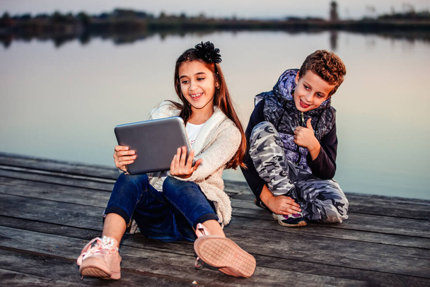 Two young cute little friends, boy and girl having fun and  making selfie for blogging on digital tablet while sitting by the lake in the evening. The boy showing gesture thumb up. Friendship - Фото, изображение