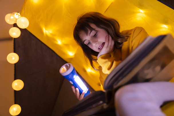 Fiatal női gyermek enciklopédiát olvas egy házi készítésű nappali sátorban fénygolyókkal. - Fotó, kép