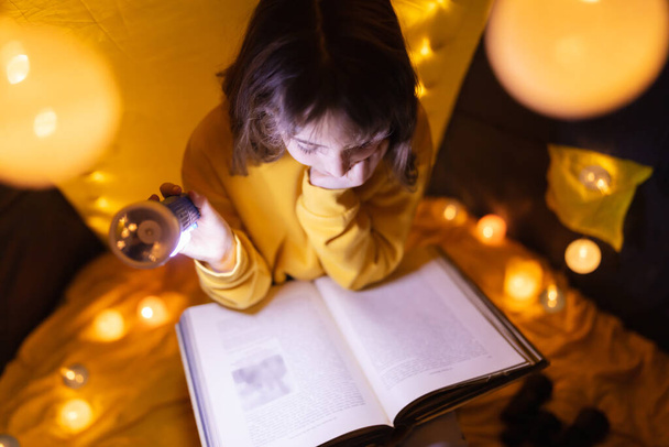 Νεαρό θηλυκό παιδί διαβάζει εγκυκλοπαίδεια σε ένα σπιτικό σαλόνι σκηνή με μπάλες φωτός. - Φωτογραφία, εικόνα
