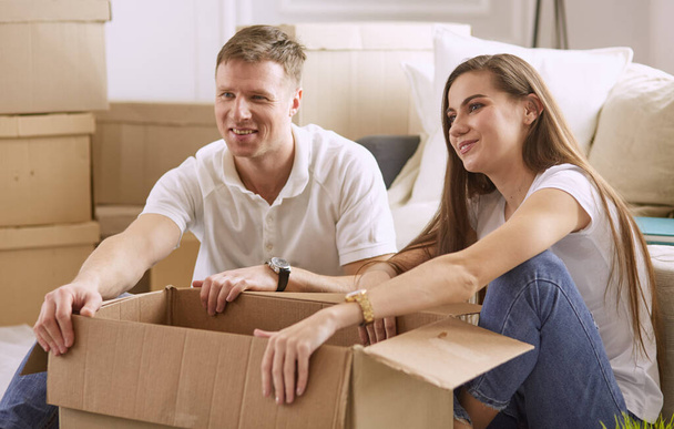Retrato de pareja feliz mirando el ordenador portátil juntos sentados en una casa nueva, rodeados de cajas
 - Foto, Imagen