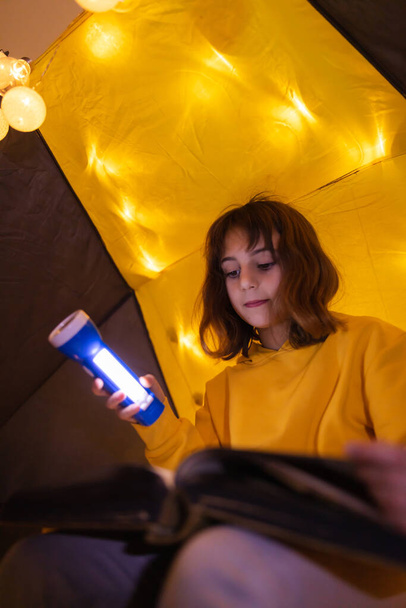 Jovem criança do sexo feminino lendo enciclopédia em uma casa feita tenda sala de estar com bolas de luz
. - Foto, Imagem