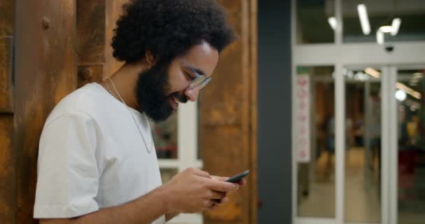 Side view of happy guy in 30s using his smartphone while standing leaned to wall. Cheerful man in glasses and white t shirt scrolling neews feed in social networks and smiling . Indoors. - Footage, Video