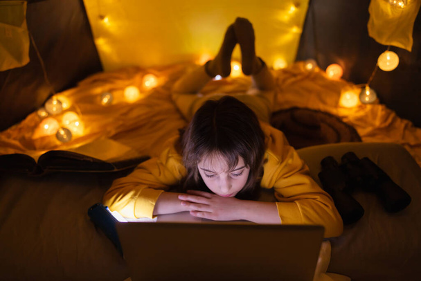 Μικρό 11χρονο κορίτσι που χρησιμοποιεί φορητό υπολογιστή κάτω από την σπιτική σκηνή της μέσα στο σαλόνι. - Φωτογραφία, εικόνα