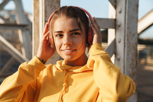 Photo of a positive optimistic pretty young fitness woman posing outdoors listening music with headphones. - Foto, imagen