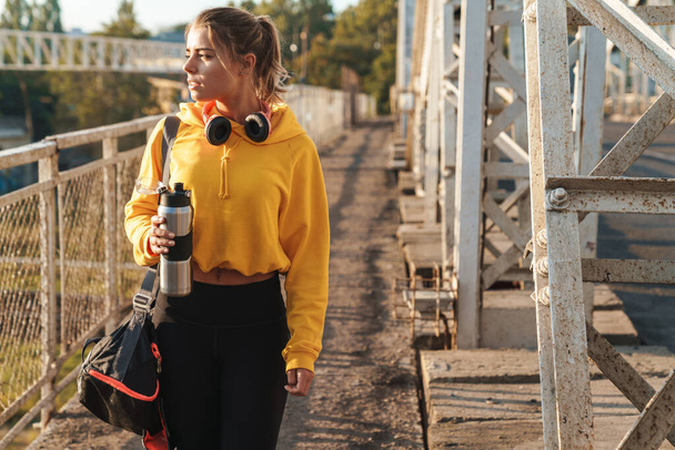 Beautiful young healthy sportsgirl wearing sportswear walking on a bridge outdoors, carrying bag, holding water bottle - Fotó, kép