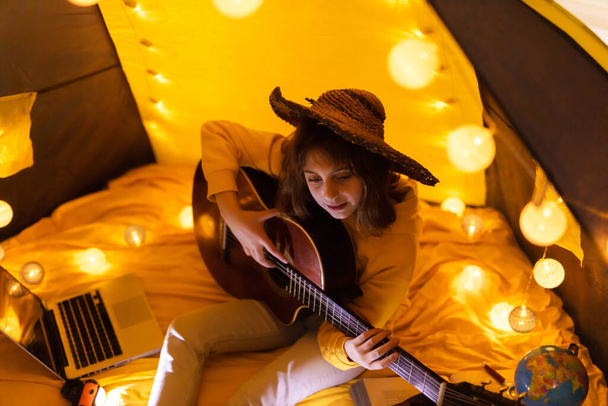 Meisje speelt akoestische gitaar onder de tent in een woonkamer. - Foto, afbeelding