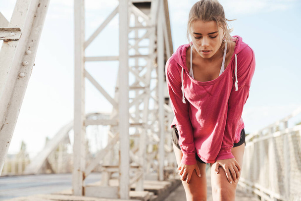 Attractive young fitness woman working out outdoors on the bridge, listening to music with earphones, resting after jogging - 写真・画像