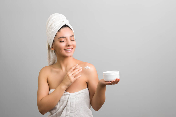 beautiful woman shrouded in towels cheerfully holding a white jar with cream and touching her body - Foto, Imagen