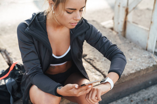 Foto de bastante joven fitness mujer sentarse al aire libre utilizando el teléfono móvil mirando reloj
. - Foto, Imagen