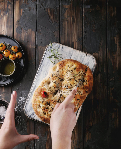 Vrouwelijke hand snijden focaccia plat brood geserveerd met gebakken tomaten, olijfolie en verse rozemarijn, met vintage servet en mes op een houten achtergrond. Bovenaanzicht - Foto, afbeelding