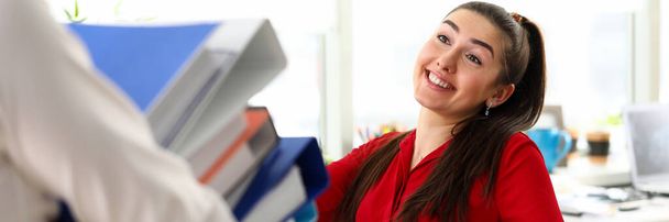 Νεαρή ευτυχισμένη γυναίκα που κάθεται στο γραφείο και χαιρετά συνάδελφο με ένα σωρό έγγραφα - Φωτογραφία, εικόνα