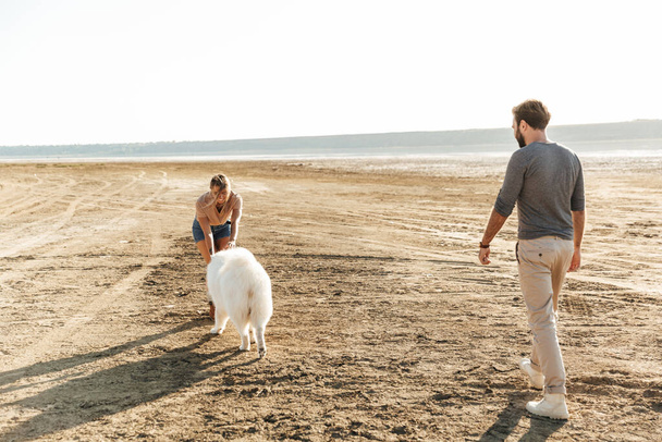 Jovem casal alegre andando na praia, brincando com seu cão
 - Foto, Imagem
