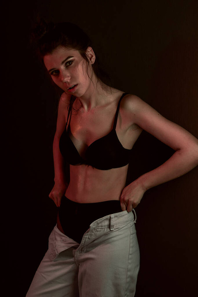 Cute young girl in black bra and pants wet skin stands posing leaning against the wall - Foto, Imagen