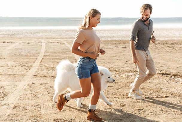 Изображение удивительной довольной молодой, удивительной любящей пары с собакой на пляже
. - Фото, изображение