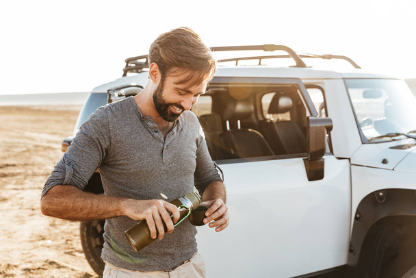 Image of positive handsome young man outdoors at the beach near car drinking tea. - Foto, Bild