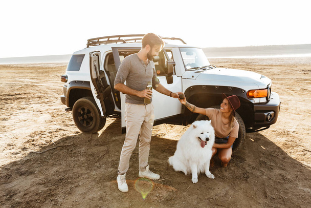 Image of positive young amazing loving couple with dog samoyed outdoors at the beach near car. - Fotó, kép