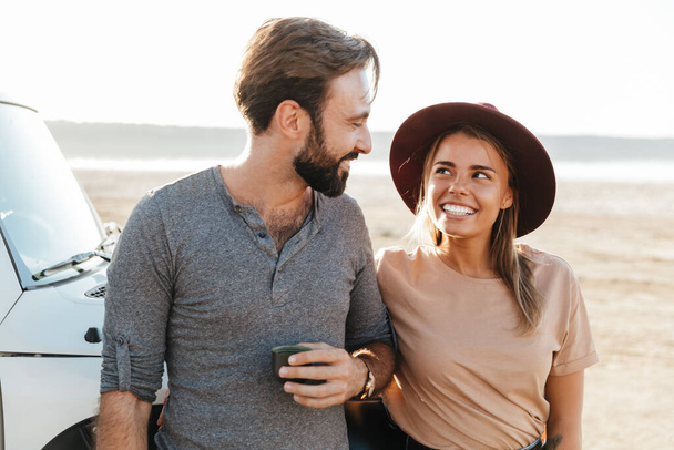 Image of happy young loving couple outdoors at the beach near car drinking tea. - Φωτογραφία, εικόνα