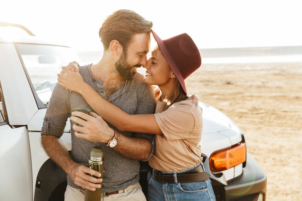 Photo of young cheerful smiling loving couple outdoors at beach near car drinking coffee. - Zdjęcie, obraz