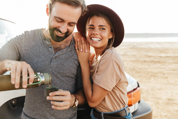 Photo of young happy loving couple outdoors at beach near car drinking coffee. - Fotoğraf, Görsel