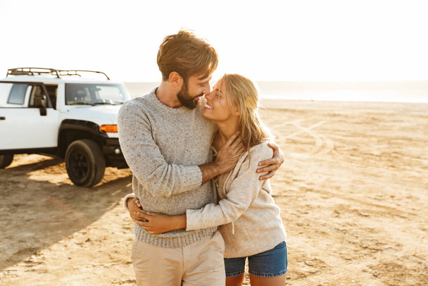 Photo of young pleased smiling happy loving couple outdoors at beach near car hugging. - Foto, imagen