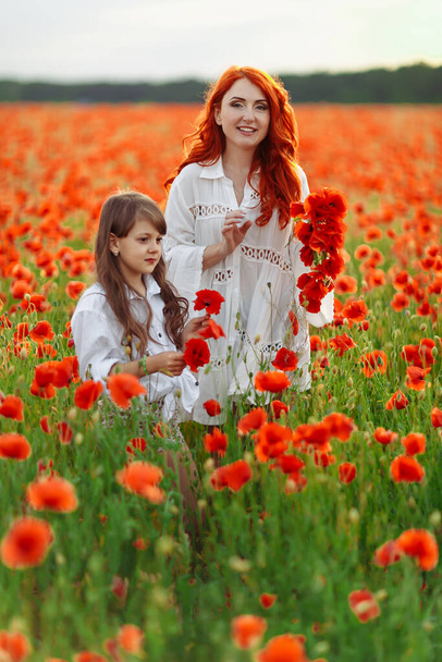 Little happy girl with redhead mother in white dresses makes wreath on poppy field at warm summer sunset - Фото, зображення