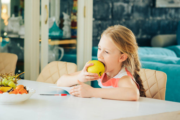 a little girl at the table with a notebook, doing homework and decided to eat a fresh Apple. - Foto, imagen