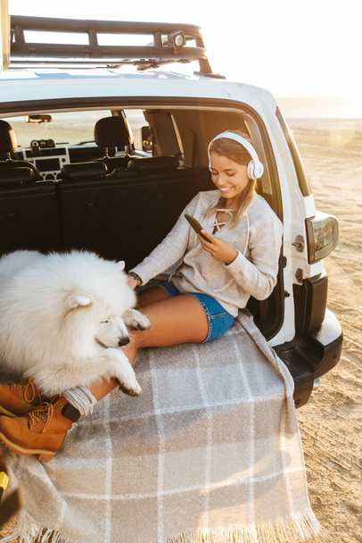 Image of smiling young woman with dog samoyed outdoors at the beach in car listening music with headphones using mobile phone. - Foto, Imagen