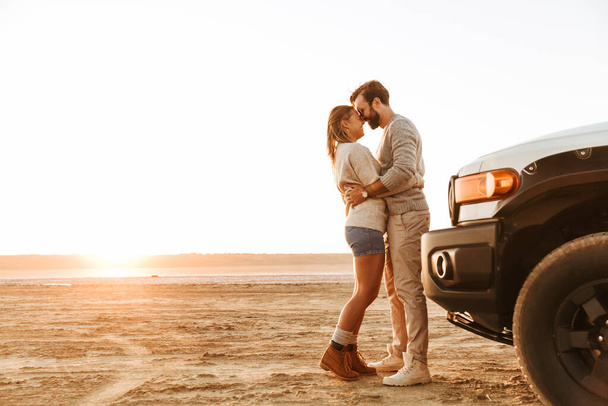 Beautiful happy young couple standing near a car at the sunny beach, hugging - Фото, зображення