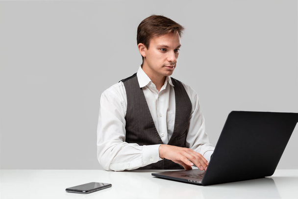 Бизнесмен в белой футболке и сером жилете, работающий на ноутбуке. Человек смотрит в камеру
 - Фото, изображение