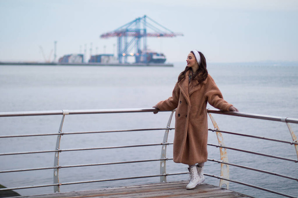 Молодая девушка в шубе улыбается на набережной, морской порт на заднем плане
 - Фото, изображение