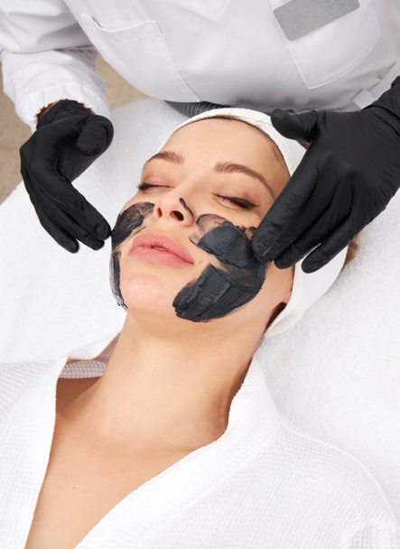 Kozmetológus fekete maszkot visel egy gyönyörű nő arcán szénhámlásra. - Fotó, kép