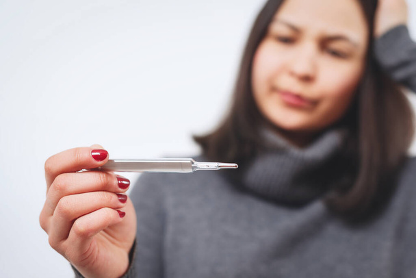 A concerned young woman in a gray sweater, holding a thermometer. Healthy lifestyle, treatment of patients with diseases, the concept of the cold season. The layout of copy space - Fotoğraf, Görsel