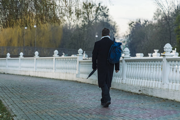 mężczyzna w czarnym płaszczu powrót do kamery spacery aleja droga dzielnica park przestrzeń wzdłuż białego ogrodzenia architektura obiekt depresja nastrój jesień sezon czas - Zdjęcie, obraz