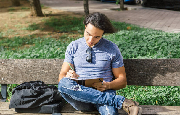 Atraktivní mladý chlapec s modrou košilí píše uvolněně ve svém deníku, sedí na lavičce v parku za slunečného jarního dne - Fotografie, Obrázek