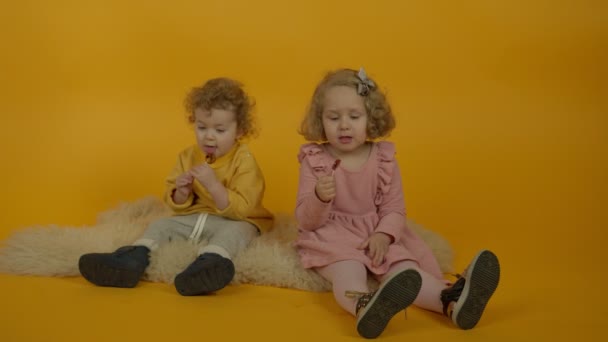 Twee krullende kinderen zitten op tapijt en eten lolly 's - Video