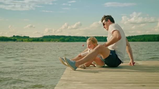 handsome man funny daughter sit on river pier at sunlight - Footage, Video